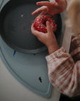 A child in a pink plaid shirt holds a red-frosted, sprinkle-topped donut over a gray plate placed on a mushie Silicone Placemat made from food-grade silicone.