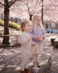 Two toddlers stand outdoors under cherry blossoms. One in cream holds a toy, while the other, wearing glasses and a lavender sweater, sips from a mushie Snack Cup on this sunny day.