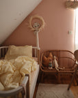 A cozy children’s bedroom with a small bed and yellow bedding, a rattan bench with stuffed toys and the mushie Stacking Rings Toy, a pink accent wall, and soft daylight through a window with white curtains.