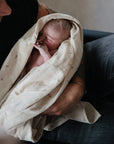 A person gently cradles a sleeping newborn wrapped in the mushie Organic Cotton Muslin Swaddle Blanket, conveying tenderness and care.