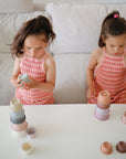 Two young girls in matching pink plaid dresses sit at a white table, stacking colorful mushie Stacking Cups Toys, deeply focused on their play. Behind them is a light sofa and a cozy space reminiscent of Rundetårn.