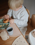 A young child in a light outfit sits at a wooden table, eating from a divided plate and sipping from mushie Dinnerware Cups. The cozy setting highlights the child's focus on their meal with safe, stylish cups by mushie.