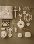 A flat lay of beige and white baby items on a green surface, including a folded muslin cloth, teethers, rattles, textured toys, a mushie Snack Cup, and small sprigs of flowers, all neatly arranged in rows.