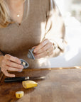 A person with long blonde hair uses the Mushie Silicone Fresh Food Feeder to store a banana piece on a wooden cutting board. Banana pieces, a knife, and the person's brown sweater are also visible.