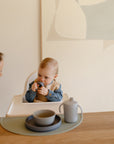 A baby in a blue shirt and bib sits in a high chair holding a toy, while an adult with long hair leans in. On the table are a plate, bowl, cup, and a mushie Silicone Placemat. Abstract art is on the wall in the background.