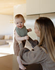 A woman supports her smiling baby, who stands on a kitchen counter wearing a mushie Muslin Bib and a brown outfit, in a bright, modern home.