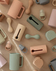 The mushie Silicone Baby Rattle Toy is displayed on a beige surface with geometric shapes, creating a modern, minimalistic flat lay pattern that highlights its refined sensory appeal.