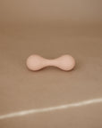 The mushie Silicone Baby Rattle Toy is a small, beige rattle made from food grade silicone with rounded ends and a slim center, shown on a plain brown surface against a soft background. Ideal for gentle sensory play.