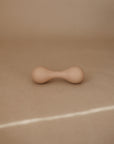 The mushie Silicone Baby Rattle Toy, in smooth beige and hourglass-shaped with rounded ends, sits on a tan surface against a brown background, offering a safe sensory experience with its food grade silicone material.