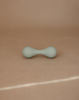 The mushie Silicone Baby Rattle Toy, featuring a small light green double-ended hourglass design, rests on a smooth beige surface with a plain background, inviting a gentle sensory experience for little hands.