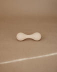 The mushie Silicone Baby Rattle Toy, featuring a smooth white hourglass shape and rounded ends, rests on a beige surface under soft lighting, providing a gentle sensory experience for babies.