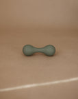 The mushie Silicone Baby Rattle Toy, small and green with rounded ends and an hourglass shape, rests on a beige surface against a matching background, providing a gentle sensory experience for babies.