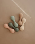 The mushie Silicone Baby Rattle Toy features five bone-shaped pieces in muted peach, sage green, dark green, beige, and light brown made from food-grade silicone, arranged on a smooth tan surface for safe sensory play.