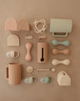 A flat lay of pastel baby items, featuring mushie’s Silicone Baby Rattle Toy along with food-grade silicone mugs, pacifiers, teething rings, soft dumbbells, rainbow toys, and a beaded teether—all arranged for a charming sensory display.
