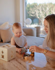 A woman and baby sit at a table in a bright living room, playing with wooden blocks and a shape sorter. The baby wears a mushie Muslin Bib as the woman smiles, gently guiding playtime fun.