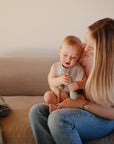 A woman with long blonde hair sits on a beige couch, smiling at her baby on her lap. The baby wears a mushie Muslin Bib and holds a light blue object, while a green cloth is draped over the couch.