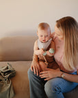 A woman sits on a beige sofa, smiling as she holds a baby playing with the mushie Silicone Baby Rattle Toy. The baby enjoys the light-colored rattle while a green cloth rests beside them on the sofa.