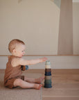 A baby in a brown outfit sits on the wooden floor, reaching for the colorful mushie Stacking Cups Toy. Neutral-toned wall art in the background adds to the cozy Danish Hygge-inspired charm.