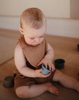A baby in a brown outfit sits on a wooden floor, playing with toy cups and wearing the mushie Muslin Bib made from soft organic cotton muslin.