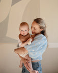 A smiling woman in a light blue floral blouse holds a baby wearing the mushie Muslin Bib. The baby faces the camera, and abstract neutral wall art decorates the background.