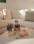 The mushie Nesting Stars Toy is arranged with pastel blocks on a white coffee table in front of a white sofa, creating a cozy, softly-lit living room perfect for Danish Hygge style and encouraging fine motor skill development.