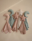 Three mushie Moon Lovey Blankets in soft pastel pink, blue, and white organic cotton are displayed on a beige surface, each featuring a plush crescent moon or star top that matches the comforter’s gentle tones.
