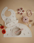 On a beige surface, the mushie Snack Cup is shown with bib, bowl and spoon, placemat, pacifiers, flower-shaped teether, and red poppy flowers. All items are in soft pastel shades with floral patterns, ideal for toddlers.