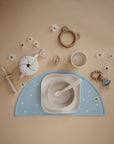 A flat lay of baby and toddler feeding essentials on a beige surface: a blue floral placemat, bowl, spoon, cup, mushie Snack Cup, pacifiers, teether, spill-proof feeding syringe, and scattered chamomile flowers.