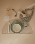 The mushie Silicone Placemat is arranged on a beige surface with a paisley bib, pacifier clip, white and green bowl, spoon, sippy cup, and feeding pacifier for safe and easy clean-ups.