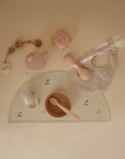 Flat lay of pastel baby items on a cream background, featuring a teapot, rattle, bib, food-grade silicone bowl with spoon, teether, and pacifier clip—all arranged on a cherry-printed mushie Silicone Placemat.