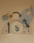 A beige baby feeding set featuring a bib, sippy cup, pacifier clip, spoon, bowl, rattle, and food-grade silicone feeder is arranged on a floral mushie Silicone Placemat against a tan background.