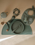 A sage green mushie Silicone Placemat is neatly arranged with baby feeding essentials—plate, spoon, sippy cup, bib, teether, pacifier with clip, and textured ball—all made from food-grade silicone.