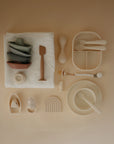 Flat lay of mushie feeding and care items in neutral tones, featuring the Silicone Baby Rattle Toy for sensory play, stackable cups, plates, bowls, a teether, and a rainbow mat—all made from food-grade silicone on a beige background.