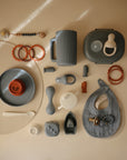 Assorted baby feeding items and teethers, including the mushie Muslin Bib, cup, plate, spoon, and silicone toys in muted shades of gray, beige, and rust are arranged neatly on a beige background.