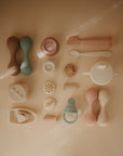 A flat lay on a beige surface features mushie's pastel Silicone Baby Rattle Toy, food grade silicone spoons, sippy cup, teether, pacifier, and accessories—neatly arranged for an enhanced sensory experience.