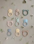Nine pastel-colored mushie silicone teethers, including the Elephant Teether, are arranged in a grid on a tan surface with small white flowers, adding charm to any baby teether collection.