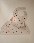 A mushie Silicone Placemat and matching pale pink food-grade silicone baby bib, both adorned with red butterfly prints, are laid flat on a neutral background. The bib includes a food catcher pocket and button fasteners.
