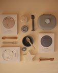 A mushie Lunchbox with built-in compartments is arranged neatly on a beige surface alongside boxed muslin cloths, silicone utensils and plates, a star-shaped teether, and stacking toys made from non-toxic plastic.