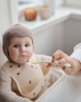 A baby in a knitted hat and butterfly bib sits in a high chair while an adult gently feeds them with a mushie Silicone Feeding Spoon. The baby gazes calmly at the camera, candles softly glowing in the background.
