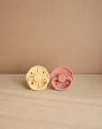 The FRIGG Moon Natural Rubber Pacifier 2-Pack by FRIGG in yellow and pink is displayed side by side on a beige surface, set against a muted light brown wall.
