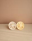 The FRIGG Moon Natural Rubber Pacifier 2-Pack by FRIGG features two soothing pacifiers—one cream, one light yellow—with crescent moon cutouts, perfect as charming essentials for any nursery.