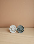 Two FRIGG Moon Natural Rubber Pacifiers—one white and one gray—are placed side by side on a light wooden surface against a beige background.