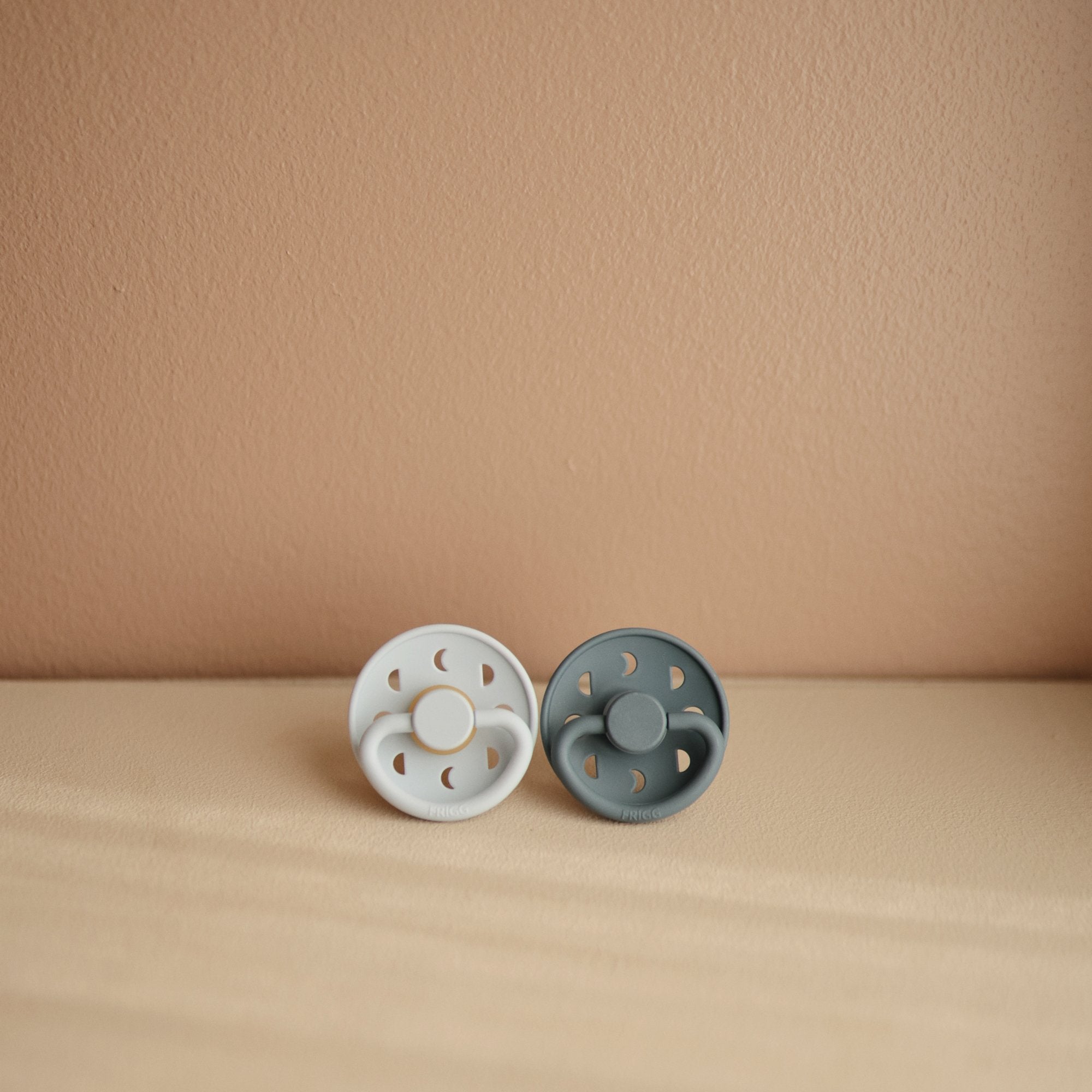 Lifestyle image of the Powder Blue and Slate Moon Silicone Frigg pacifiers.