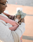 A woman in glasses carries a baby wrapped in peach. The baby, wearing a large green bow headband and using a FRIGG Andersen Fairytale Silicone Pacifier by FRIGG, looks at the camera as the woman gently supports their head.