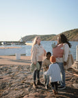 Two women wearing mushie Baby Wraps hold their babies on a stone pier by the water with two young children. Boats and hills appear in the background under a clear sky.
