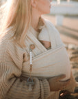 A blonde woman in a knitted sweater cradles a baby outdoors in a beige wrap carrier. A beaded clip secures the FRIGG Andersen Fairytale Silicone Pacifier by FRIGG, soft sunlight illuminating the peaceful scene.