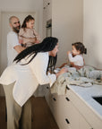Lifestyle image of a young family of four gathered around a kitchen counter with a young boy sitting on a changing pad with his mother leaning down and smiling at him. A Cambridge Blue Star Training Toothbrush and Blush Dice Press toy are laid by the baby.