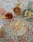 A floral tablecloth displays mushie Silicone Feeding Spoons, a bowl of cereal rings with fruit, sliced bananas and apples on plates, a cup, and a sippy cup.
