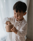A young child indoors by a softly lit window with sheer curtains reaches into a beige mushie Snack Cup while wearing a white shirt.