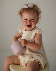 A smiling toddler with curly hair and a white bow sits on a chair, wearing a cream outfit and holding the mushie Snack Cup—a pink, spill-proof silicone cup—against a plain background.